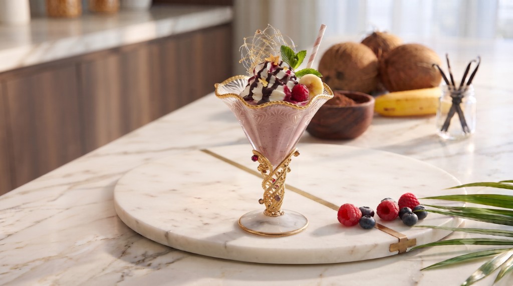 A pink berry milkshake in an ornate glass, topped with whipped cream, chocolate drizzle, banana, berries, and mint, on a marble board